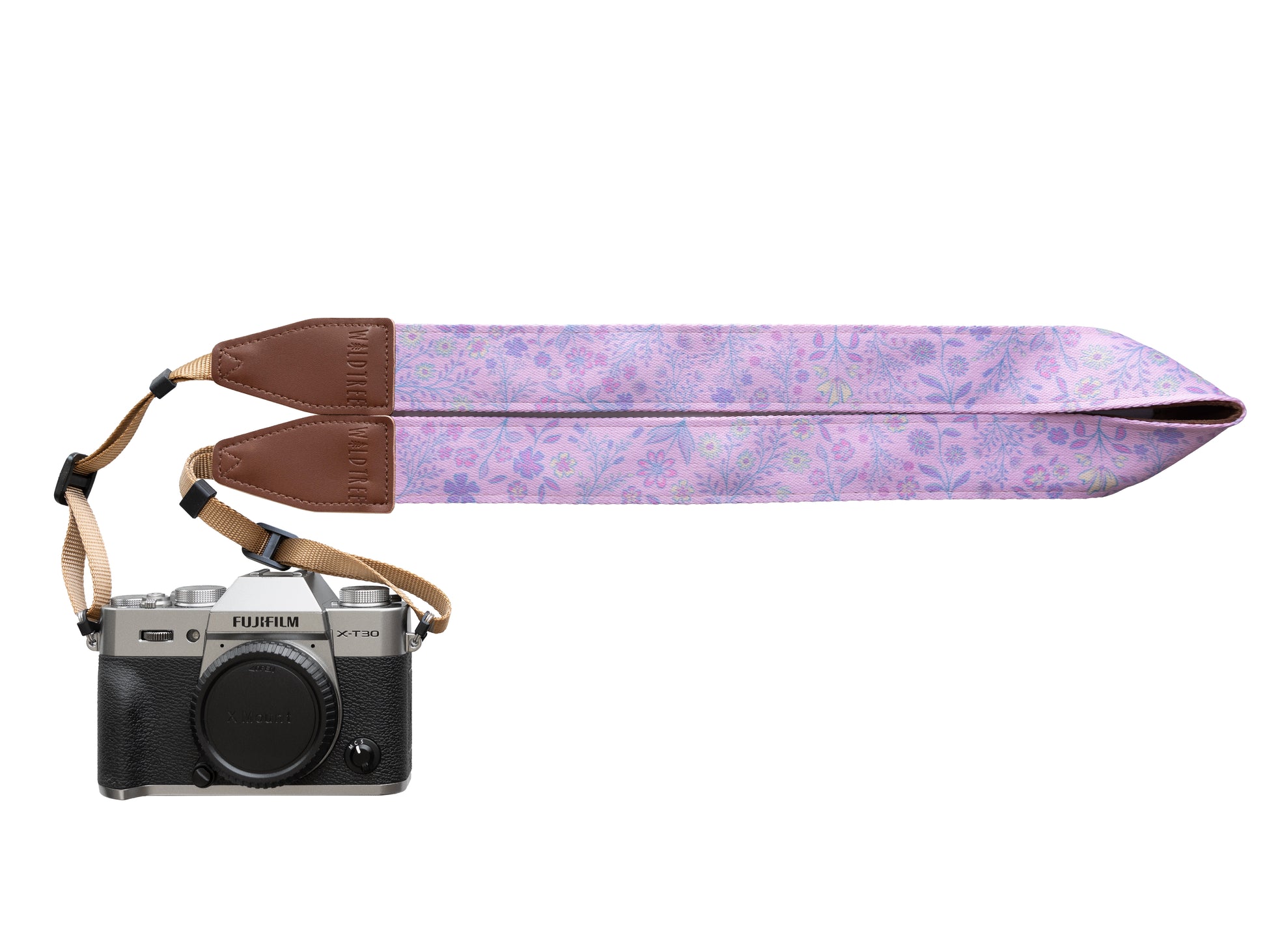 A professional studio product shot showing a Fujifilm X-T30 mirrorless camera equipped with a "Dainty Wildflower" patterned camera strap in pink. The strap features a light pink fabric with a delicate, repeating floral print, accented by brown leather attachment points and tan nylon webbing. The camera is shown from the front with a body cap attached.