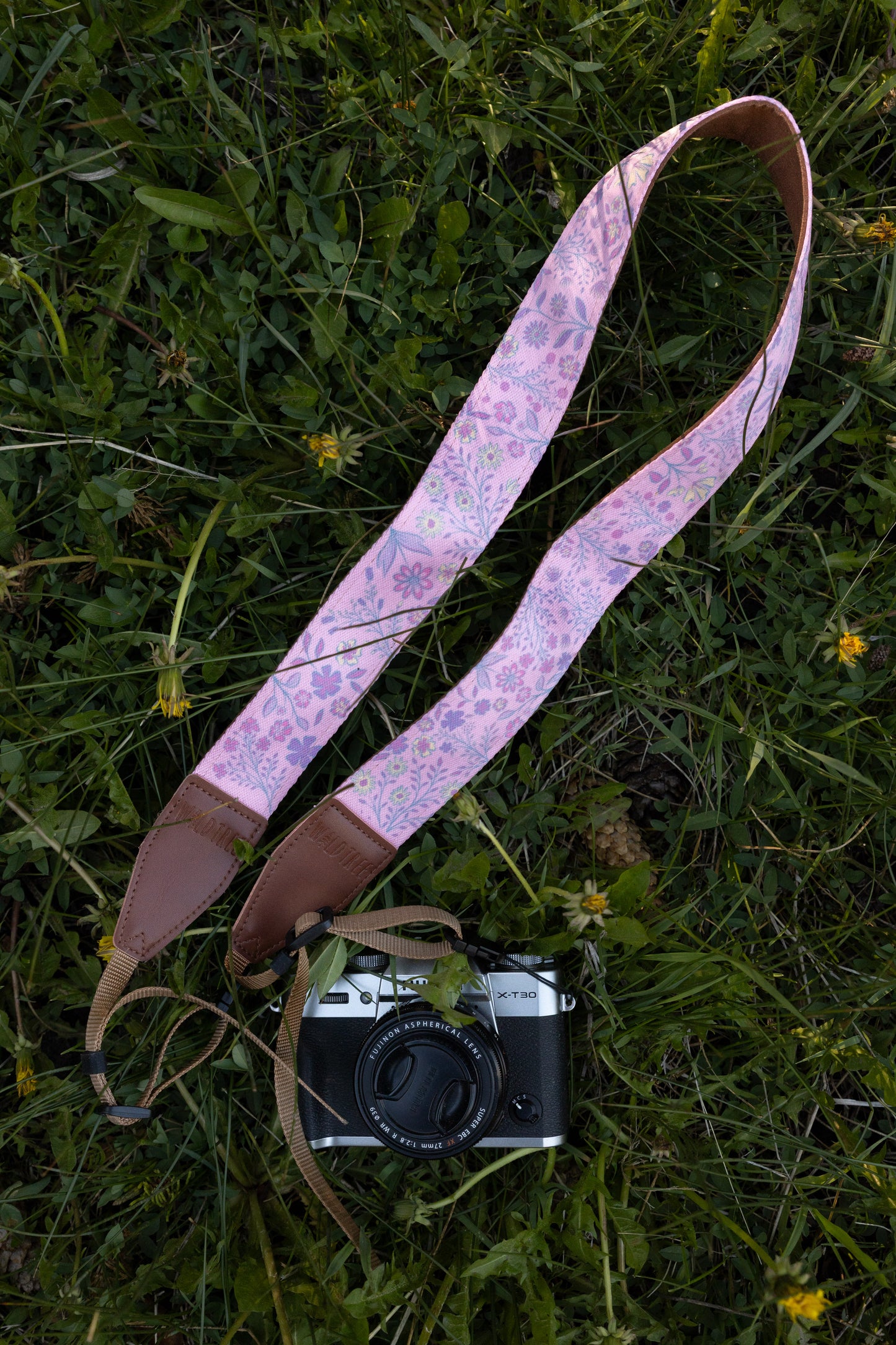 showcasing a camera strap with a vibrant pink background and a bold purple, blue, and yellow wildflower pattern, attached to a vintage silver mirrorless camera resting in green grass.