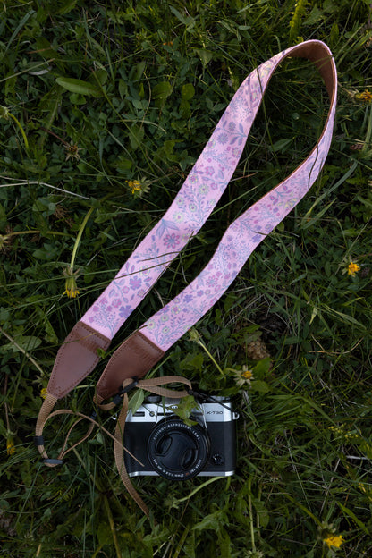 showcasing a camera strap with a vibrant pink background and a bold purple, blue, and yellow wildflower pattern, attached to a vintage silver mirrorless camera resting in green grass.
