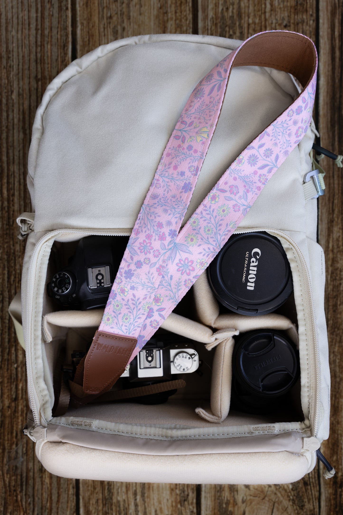 showcasing the vibrant pink wildflower print on the front and solid brown backing, positioned alongside multiple Canon camera lenses in a cream-colored (off-white) canvas camera backpack.