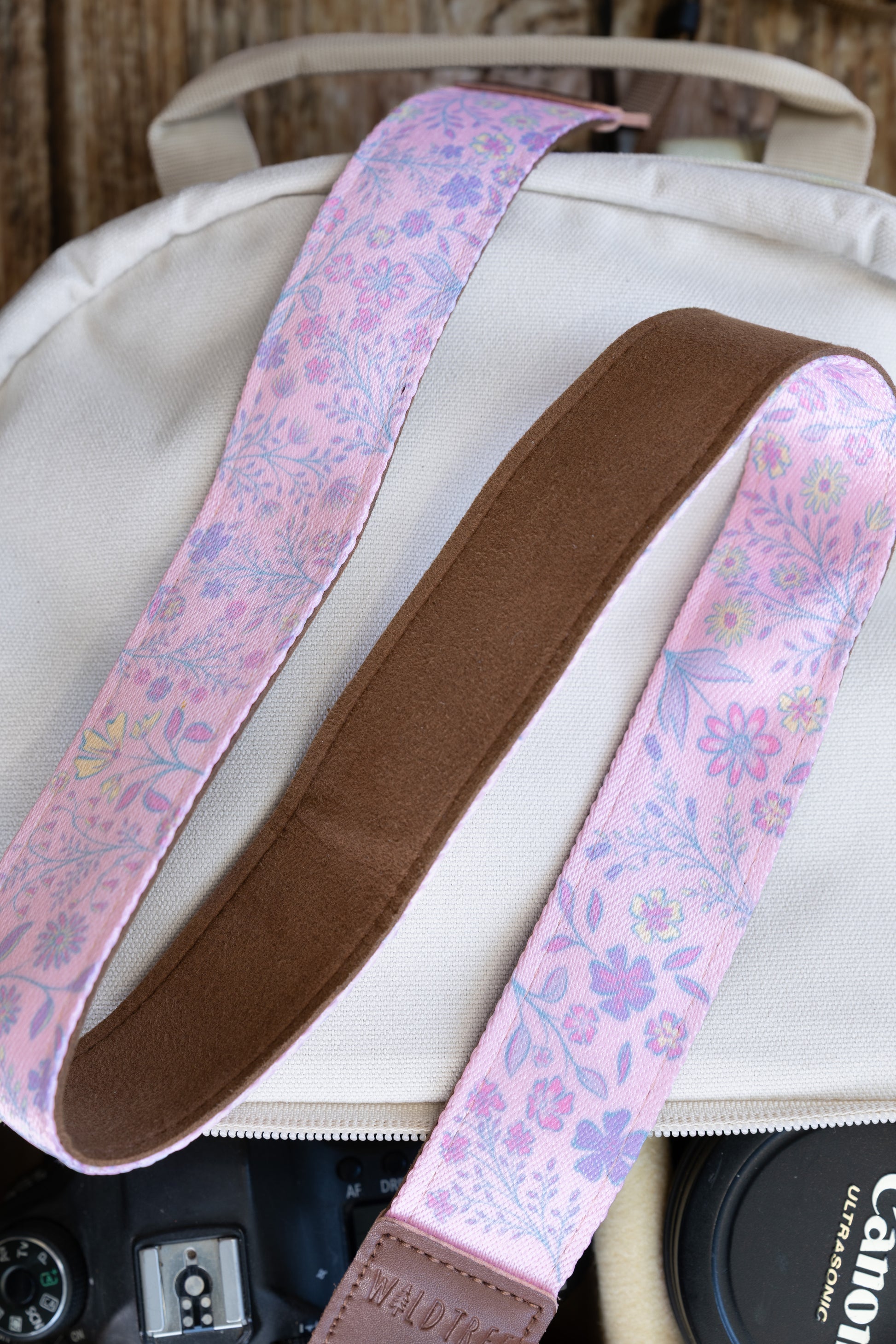 showcasing the vibrant pink wildflower print on the front and solid brown backing, positioned alongside multiple Canon camera lenses in a cream-colored (off-white) canvas camera backpack.