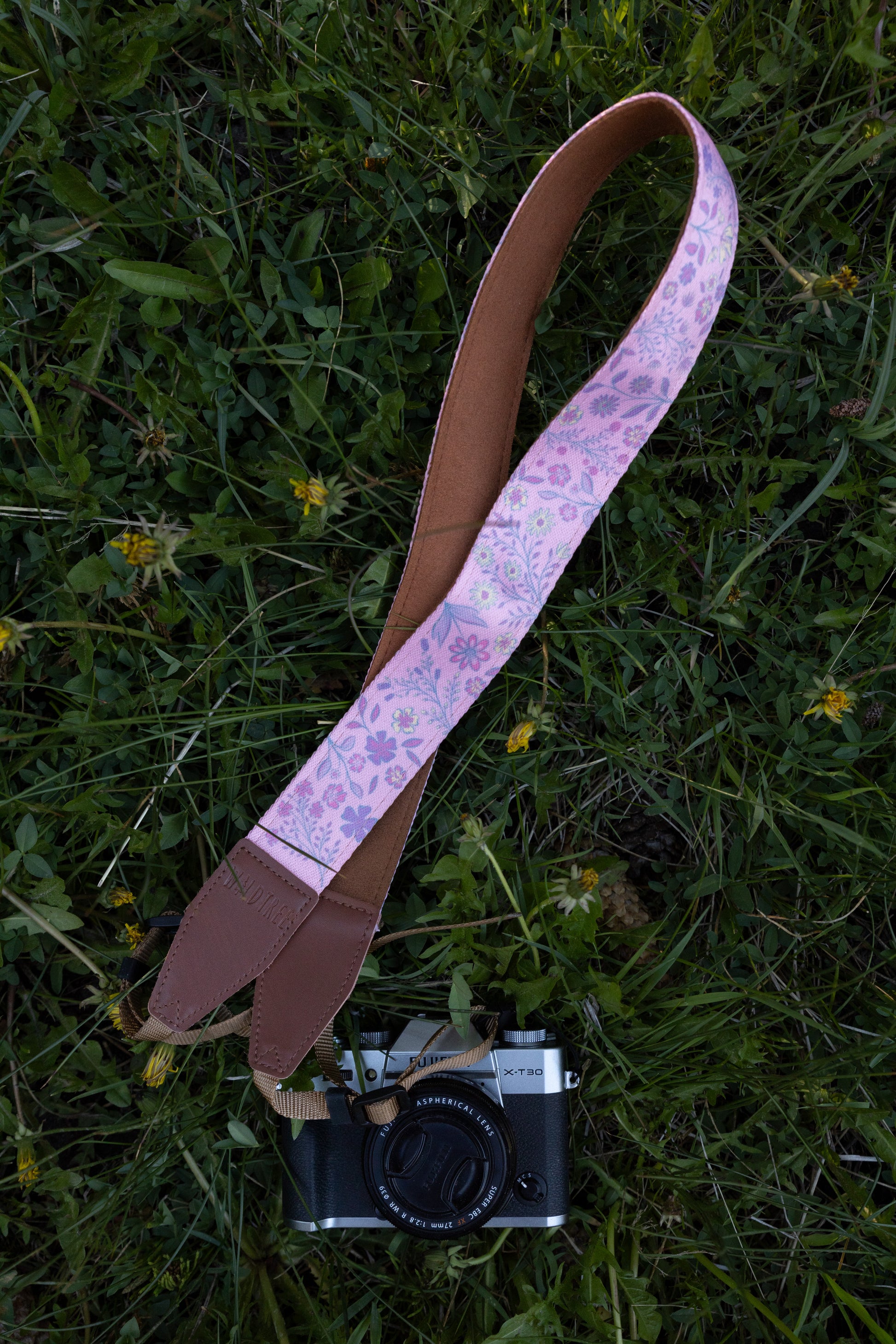 flat lay product shot showing a vibrant pink camera strap adorned with a purple, blue, and yellow wildflower pattern, laid out diagonally next to a pinecone on green grass.