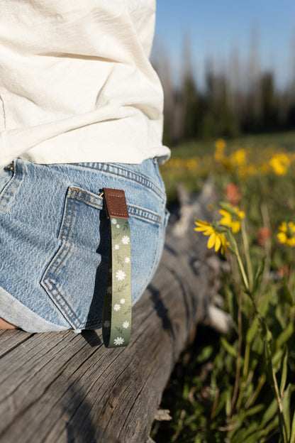 A person sitting outdoors on a log, wearing denim shorts and a white shirt. The green daisy-patterned wristlet keychain hangs from a belt loop on the back pocket of the shorts. Yellow wildflowers are visible in the blurry background.