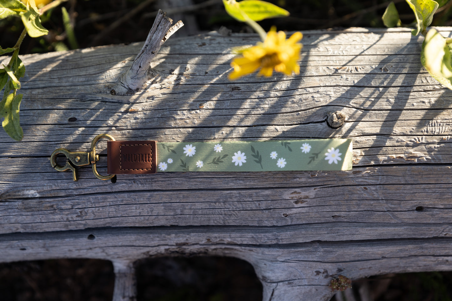 The green daisy-patterned wristlet keychain lying horizontally on a gray log. The image shows the full length of the fabric strap, the brown leather tag, and the brass lobster claw clasp. A yellow flower in the upper background is blurred.