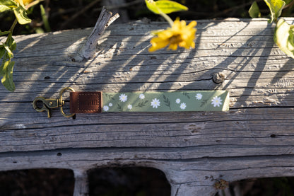 The green daisy-patterned wristlet keychain lying horizontally on a gray log. The image shows the full length of the fabric strap, the brown leather tag, and the brass lobster claw clasp. A yellow flower in the upper background is blurred.