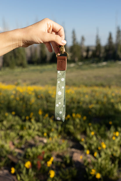 A hand holding the green daisy-patterned wristlet keychain up in front of a sunny, blurred outdoor landscape of green grass, yellow wildflowers, and trees in the distance. The wristlet has a brown leather end and a brass clasp.