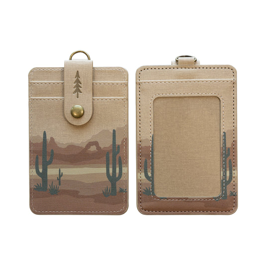 Front and back of the Desert ID badge holder, featuring a sun-washed desert landscape with layered mountain ridges and saguaro cacti, a tan top panel with an embossed pine tree logo, and a clear card window on the back.