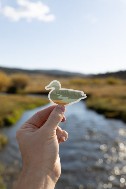 Hand holding out duck sticker in front of river and valley