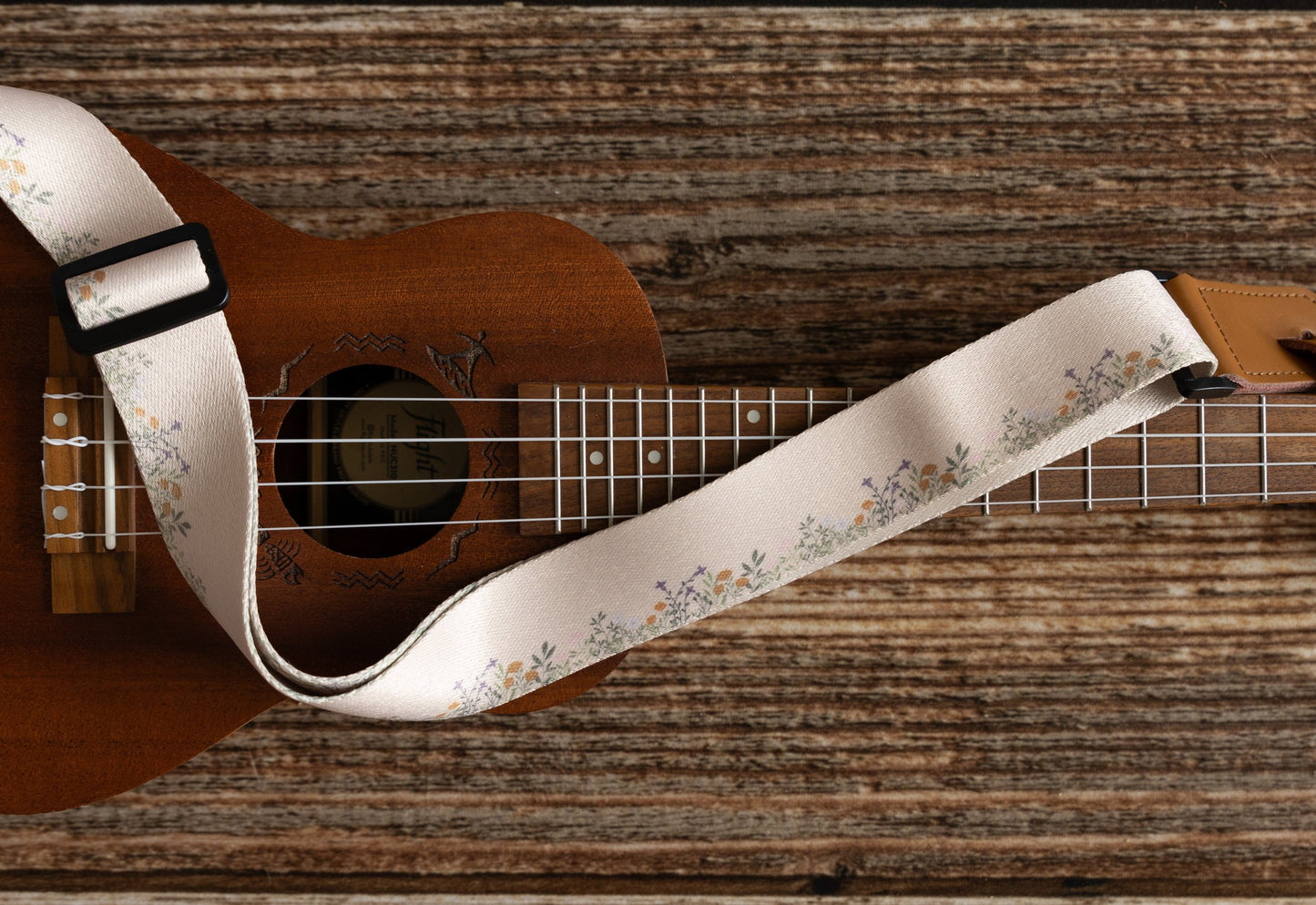 Cute tan floral strap printed with Little colored flowers across the length of the strap attached to Ukulele lying on wood floor