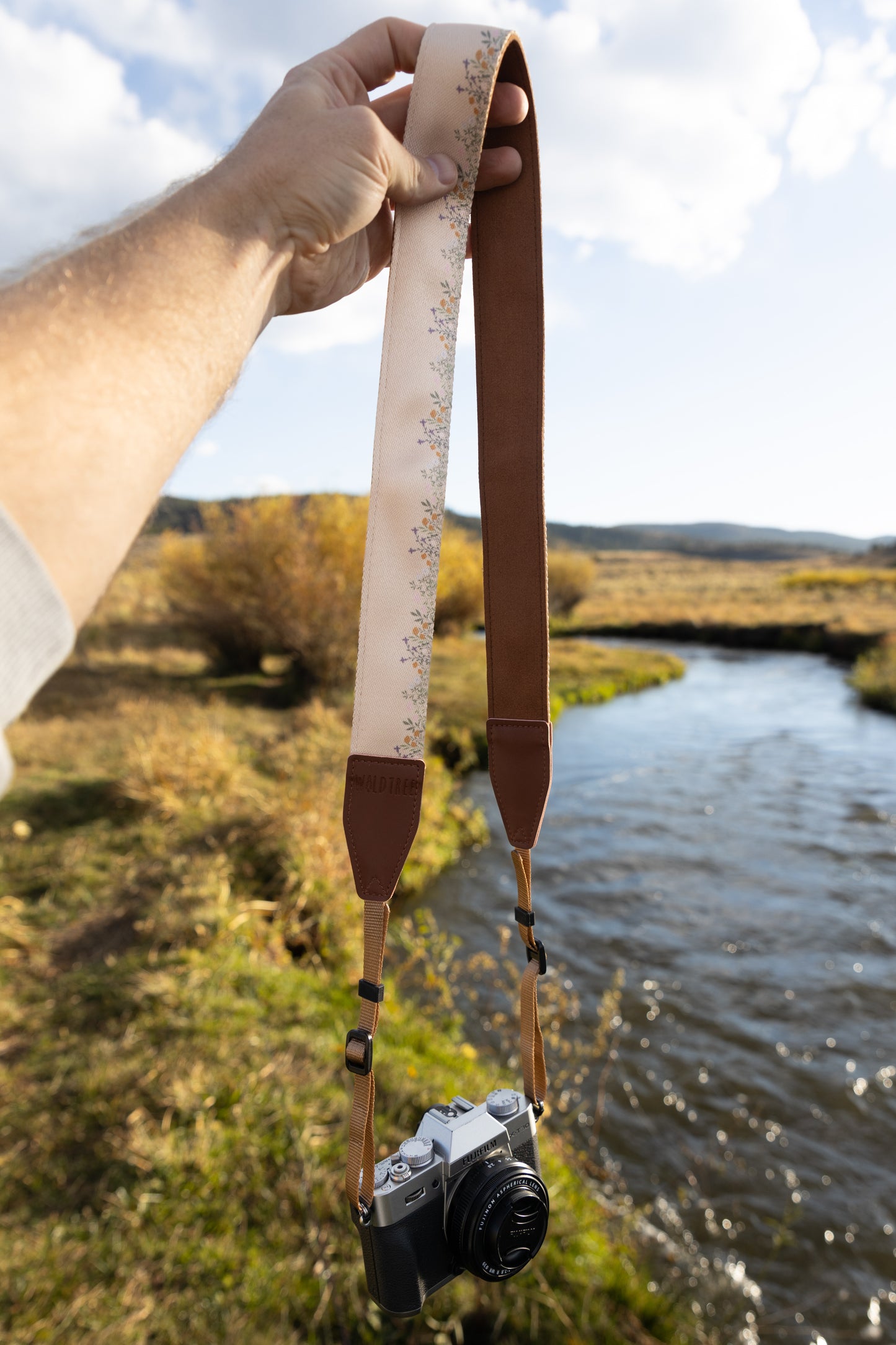 Hand holding up Flower Field Tan Camera strap. Tan strap printed with tiny little wildflowers across the length of the strap. Connected to camera 