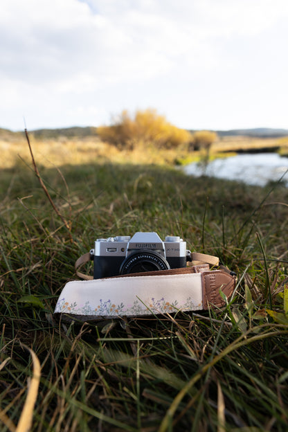 Flower Field Tan Camera strap. Tan strap printed with tiny little wildflowers across the length of the strap attached to camera lying in field of grass