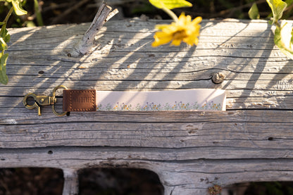 The tan wristlet keychain with a small flower border design is laid horizontally on a weathered, gray log. The brown leather tag with the "WILDTREE" logo and the brass clasp are visible on the left side of the wristlet. A single yellow flower is blurred in the upper-right area.