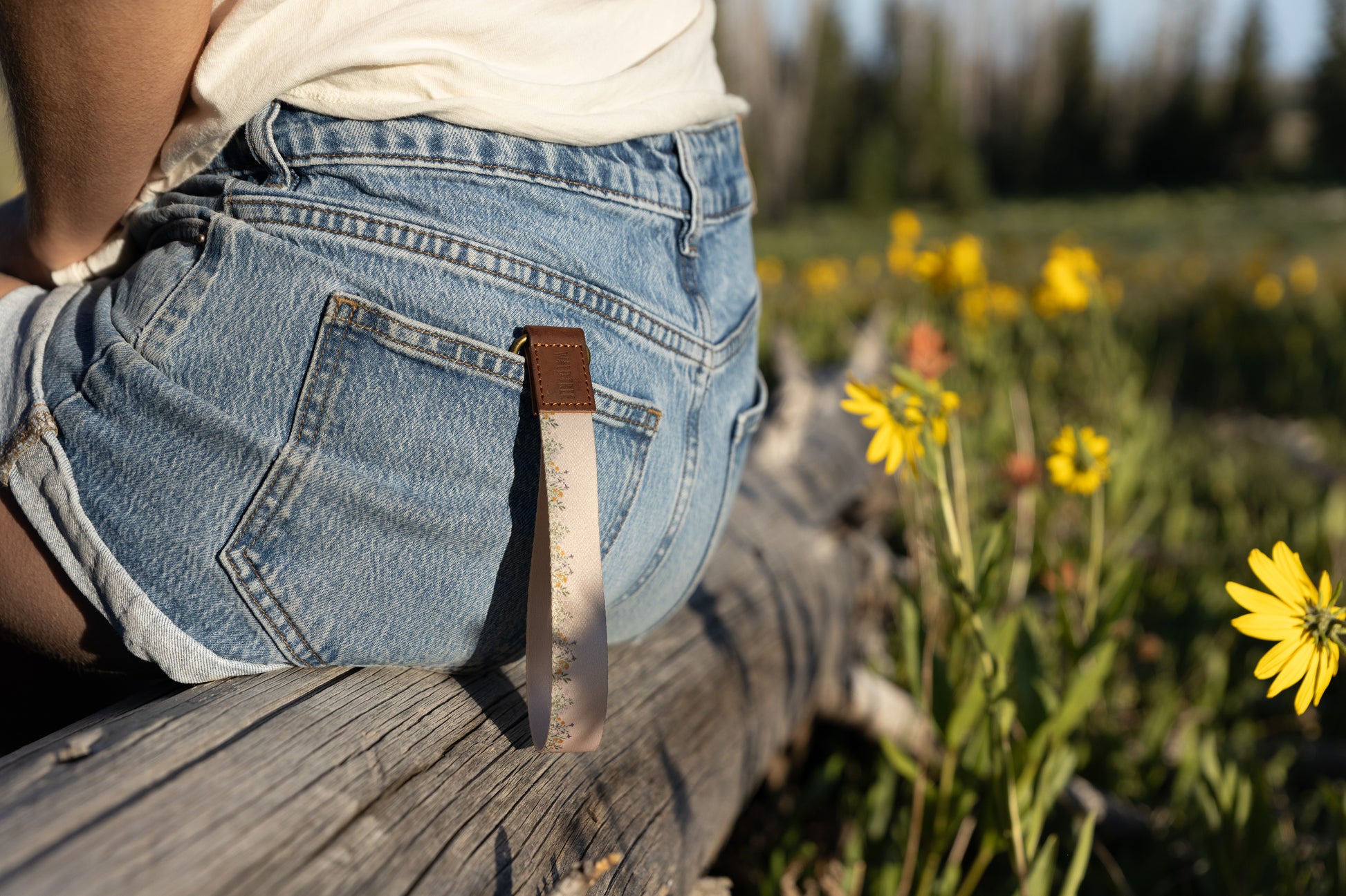 A person sitting outdoors on a weathered log, wearing denim shorts and a white top. A tan wristlet keychain with a delicate flower pattern and a brown leather end hangs from a belt loop on the back pocket of the shorts. Yellow wildflowers and green foliage are in the blurry foreground and background.