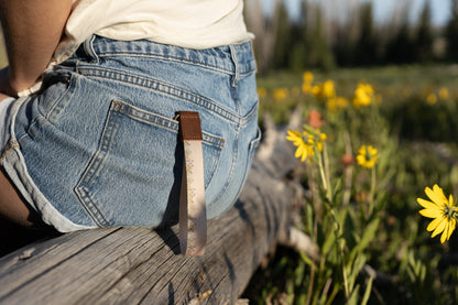 A person sitting outdoors on a weathered log, wearing denim shorts and a white top. A tan wristlet keychain with a delicate flower pattern and a brown leather end hangs from a belt loop on the back pocket of the shorts. Yellow wildflowers and green foliage are in the blurry foreground and background.