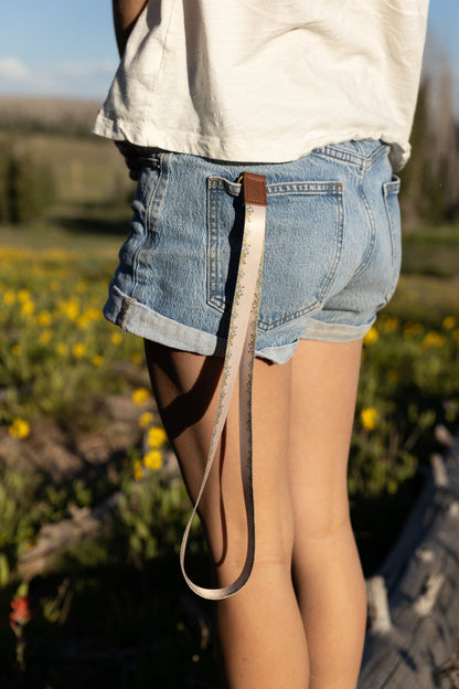 A person is standing in a field of yellow wildflowers, wearing denim shorts. The long, tan Flower Field Neck Lanyard is clipped to a belt loop on the back of their shorts, hanging down to mid-thigh. A loop of the lanyard fabric is visible at the bottom.