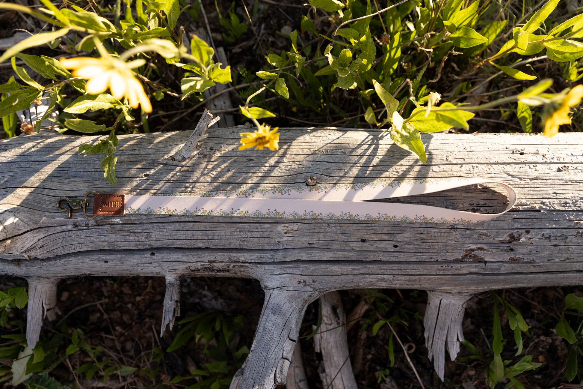 The long, tan Flower Field Neck Lanyard is laid horizontally on a weathered, gray log surrounded by green foliage and yellow wildflowers. The lanyard shows the border pattern of small yellow and green flowers, the brown leather tag, and the brass clasp.