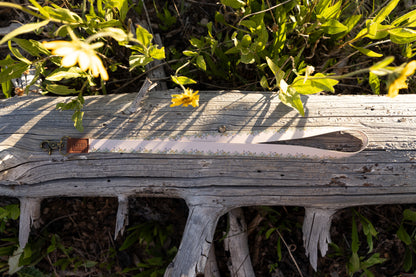 The long, tan Flower Field Neck Lanyard is laid horizontally on a weathered, gray log surrounded by green foliage and yellow wildflowers. The lanyard shows the border pattern of small yellow and green flowers, the brown leather tag, and the brass clasp.