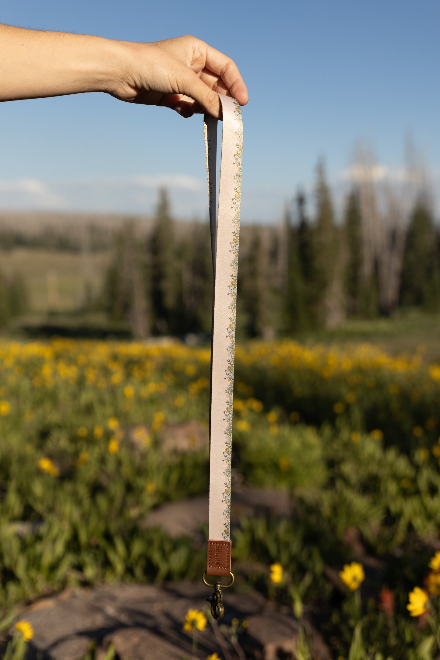 A person's hand holds a long, tan Flower Field Neck Lanyard vertically against a bright blue sky and a blurred outdoor landscape of green grass and yellow wildflowers. The lanyard has a delicate border of small yellow and green flowers, a brown leather end, and a brass clasp.
