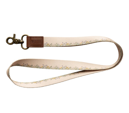 A "Wildtree flower field neck lanyard" is displayed against a stark white background. The lanyard features a tan fabric strap adorned with a delicate pattern of small yellow and green flowers and leaves running along its length.