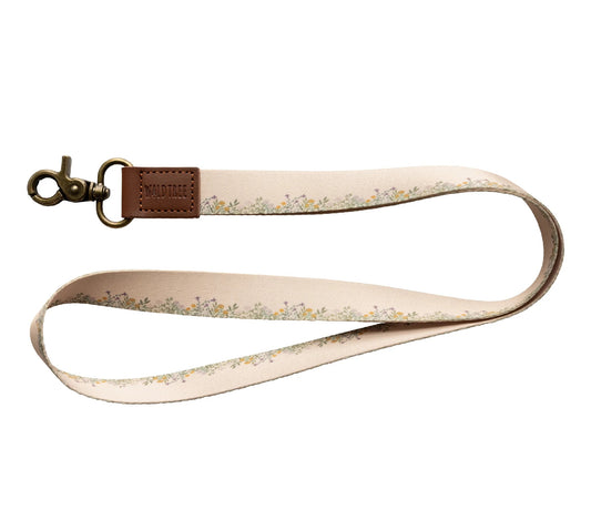 A "Wildtree flower field neck lanyard" is displayed against a stark white background. The lanyard features a tan fabric strap adorned with a delicate pattern of small yellow and green flowers and leaves running along its length.