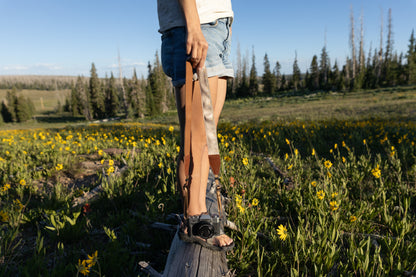 A person wearing jean shorts and a white shirt stands on a weathered tree stump in a bright, grassy field of yellow wildflowers and evergreen trees. They hold a brown and beige camera strap with a subtle floral design, which is attached to a vintage-style black and silver camera resting on the stump.