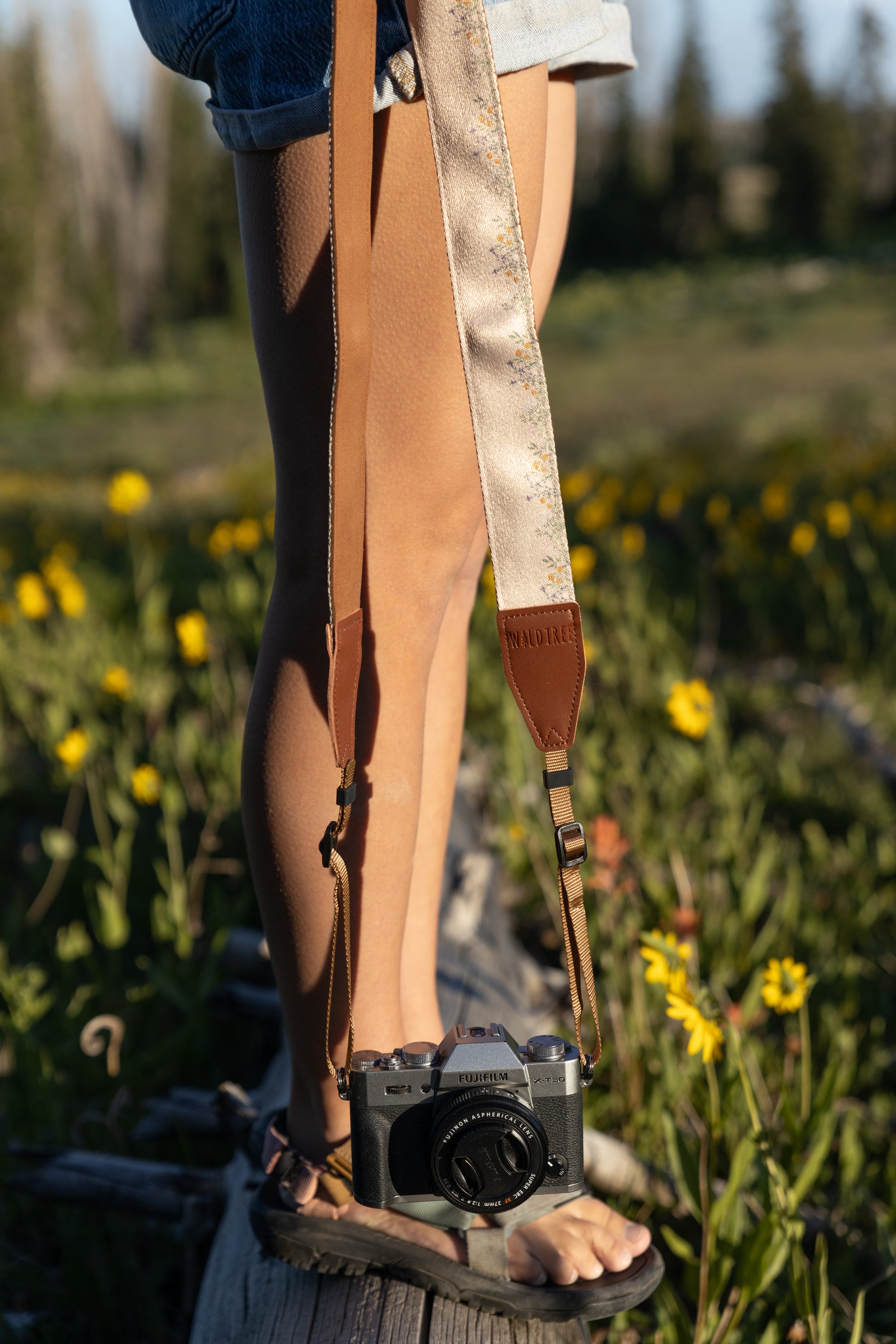 Close-up shot showing a person's legs, feet in sandals, and a Wildtree Outdoor camera strap hanging down. The strap is a two-tone design with brown leather and a textured, light beige fabric featuring a subtle, gold-and-grey floral pattern. It is attached to a vintage-style black and silver camera, all set against a blurry background of yellow wildflowers and green grass.