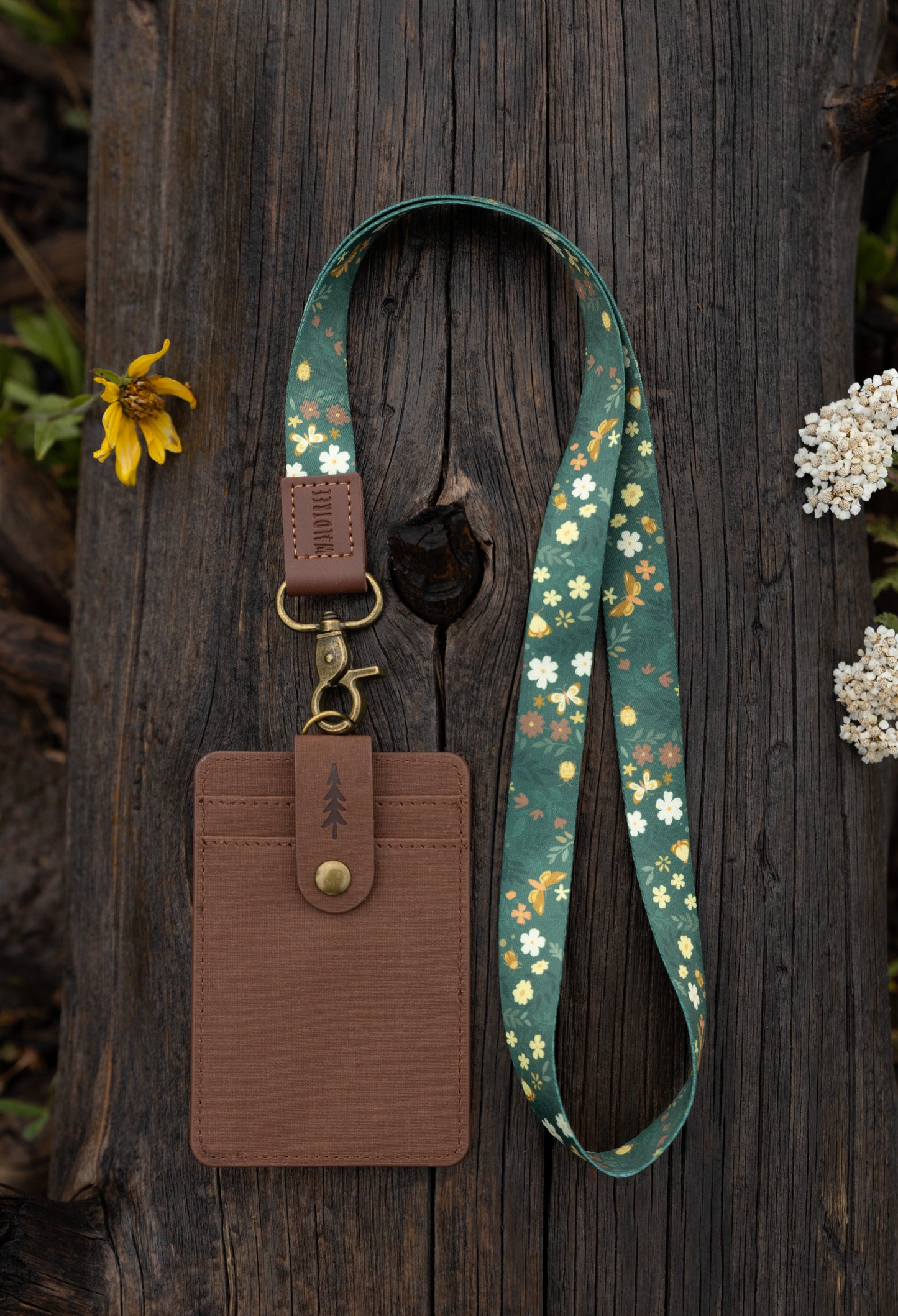 The Flutter & Fern neck lanyard laid out on a wooden log, showing the full dark green strap printed with white daisies, small brown flowers, and gold butterflies, the brown leather "WILDTREE" tab, and the bronze swivel clasp.