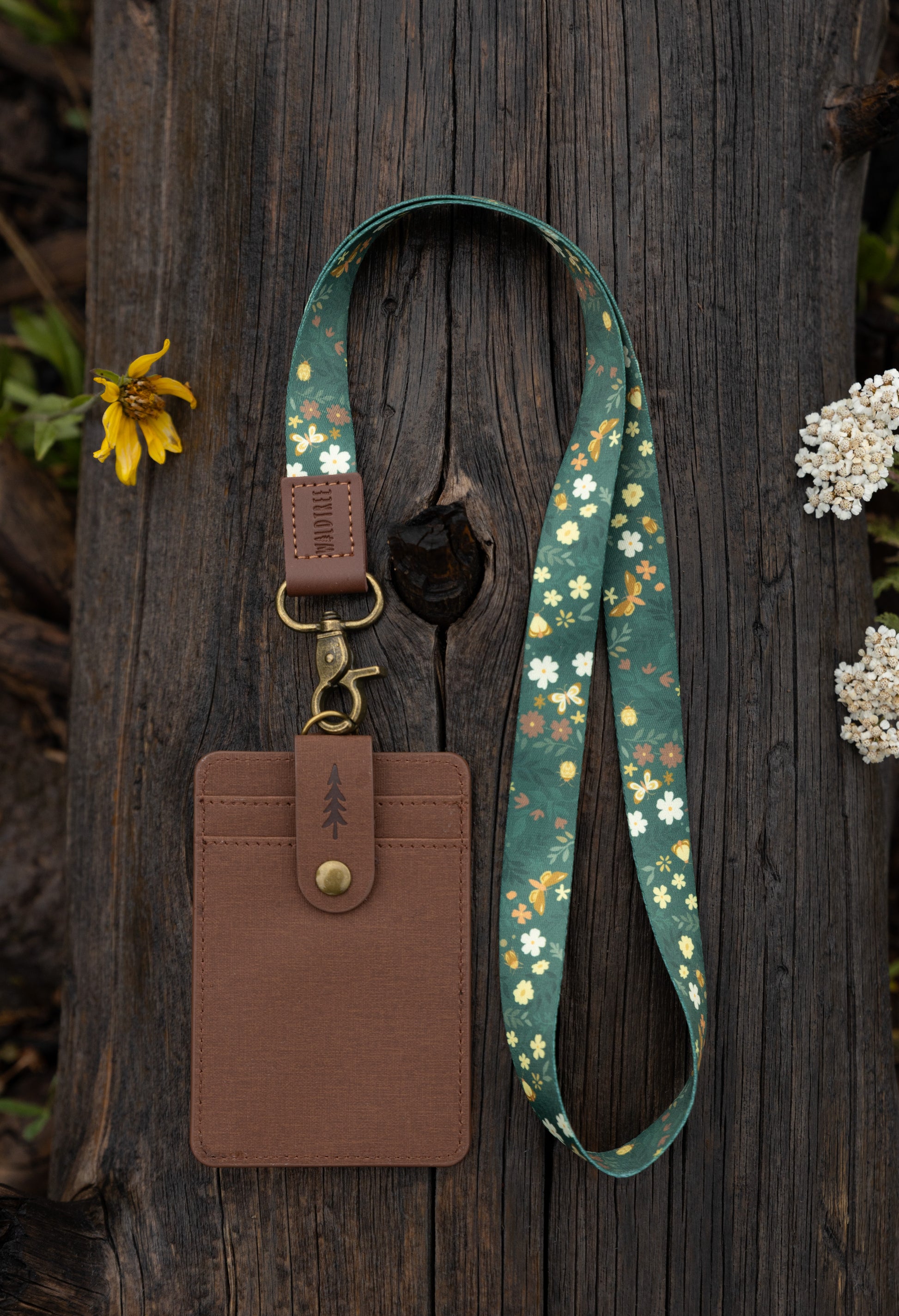 The Flutter & Fern neck lanyard laid out on a wooden log, showing the full dark green strap printed with white daisies, small brown flowers, and gold butterflies, the brown leather "WILDTREE" tab, and the bronze swivel clasp.