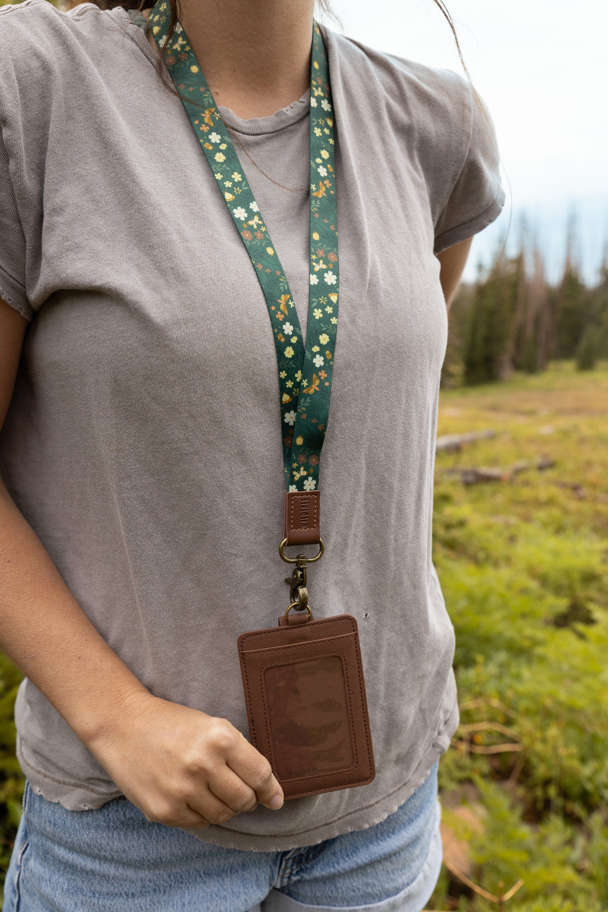 A woman wearing a gray t-shirt models the Flutter & Fern neck lanyard, its dark green strap printed with white flowers and gold butterflies visible in a close-up of the chest area, ending in a brown leather tab, bronze clasp, and brown card holder held in her hand.