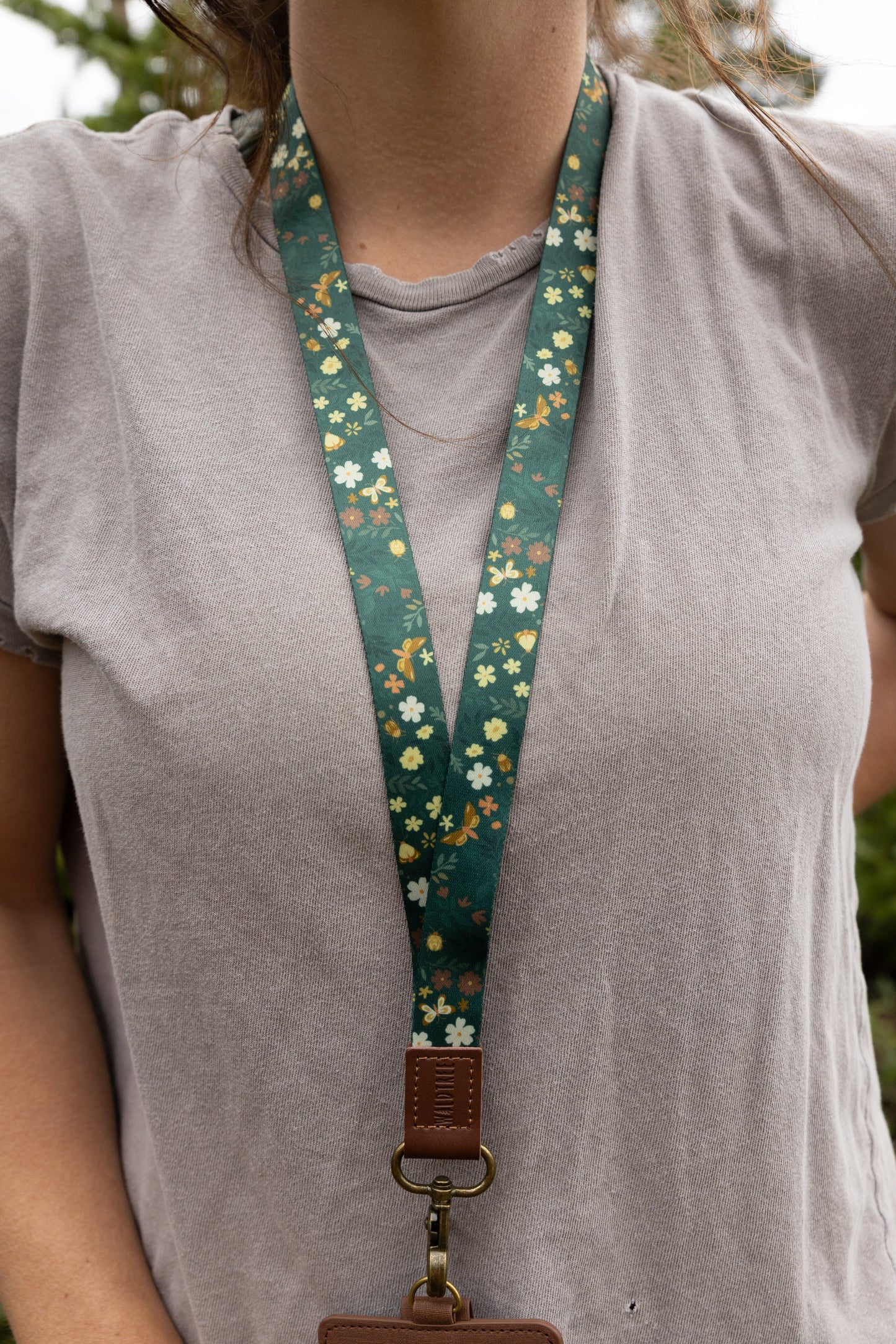 Close-up of the Flutter & Fern neck lanyard around a woman's neck, showing the dark green fabric strap patterned with white daisies, small florals, and gold butterflies forming a V shape against her gray t-shirt.