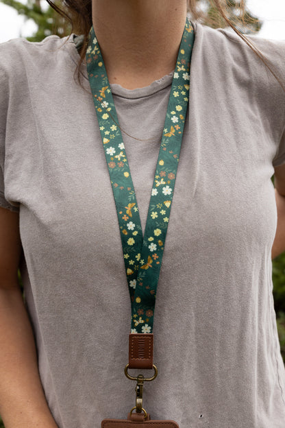 Close-up of the Flutter & Fern neck lanyard around a woman's neck, showing the dark green fabric strap patterned with white daisies, small florals, and gold butterflies forming a V shape against her gray t-shirt.