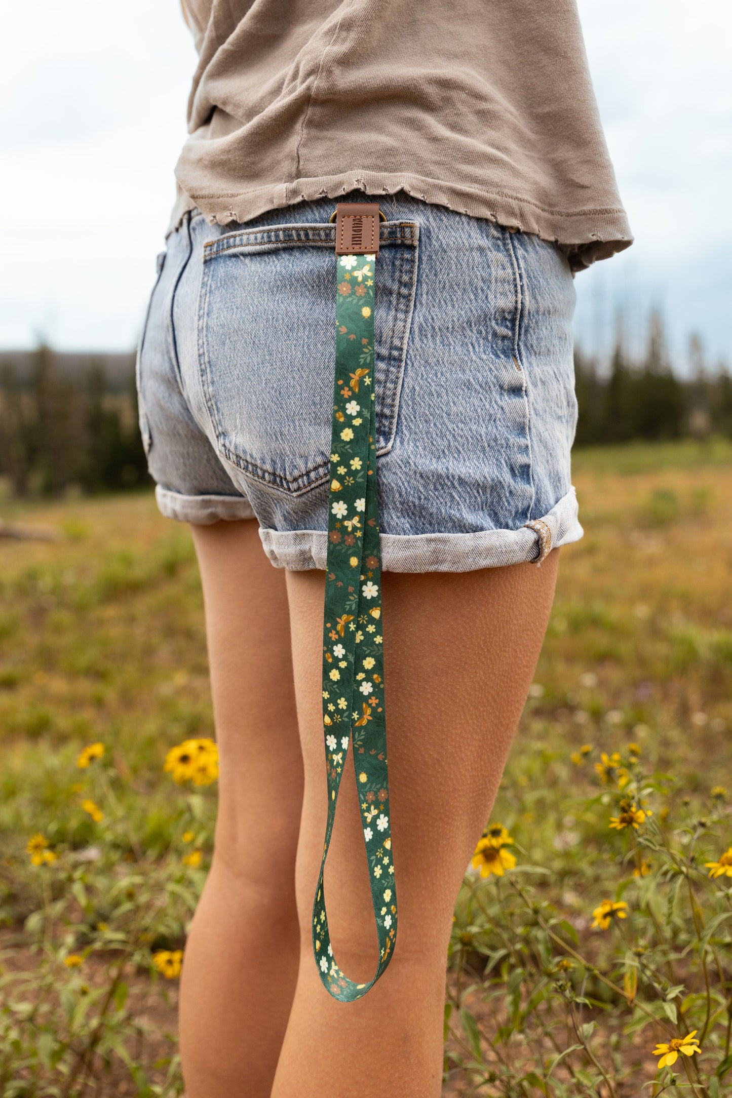 A woman in denim shorts stands in a wildflower meadow with the Flutter & Fern lanyard clipped to her back belt loop, the dark green floral and butterfly print strap hanging down against her leg.
