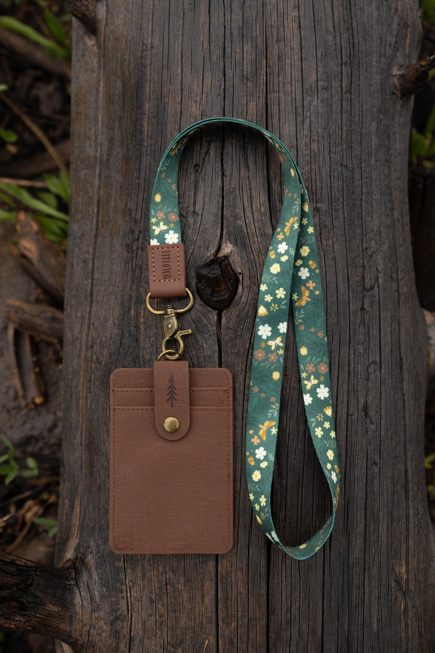 The Flutter & Fern neck lanyard laid out on a wooden log, showing the full dark green strap printed with white daisies, small brown flowers, and gold butterflies, the brown leather "WILDTREE" tab, and the bronze swivel clasp.