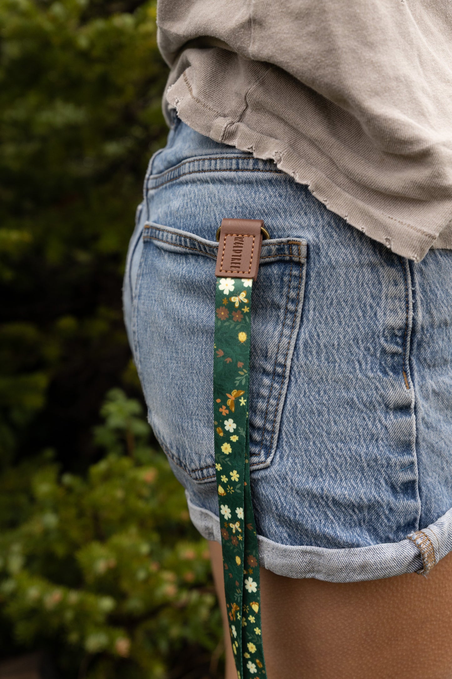 A person's hand holds a green floral neck lanyard vertically, showing its dark green fabric printed with white daisies, small brown and yellow flowers, and gold butterflies. A brown vegan-leather tab reading "WILDTREE" connects to a bronze swivel clasp holding a brown card holder with a clear ID window, set against a blurred pine forest and meadow background.