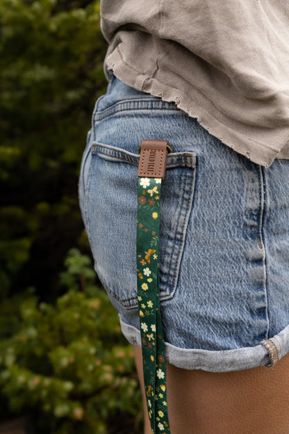 A person's hand holds a green floral neck lanyard vertically, showing its dark green fabric printed with white daisies, small brown and yellow flowers, and gold butterflies. A brown vegan-leather tab reading "WILDTREE" connects to a bronze swivel clasp holding a brown card holder with a clear ID window, set against a blurred pine forest and meadow background.