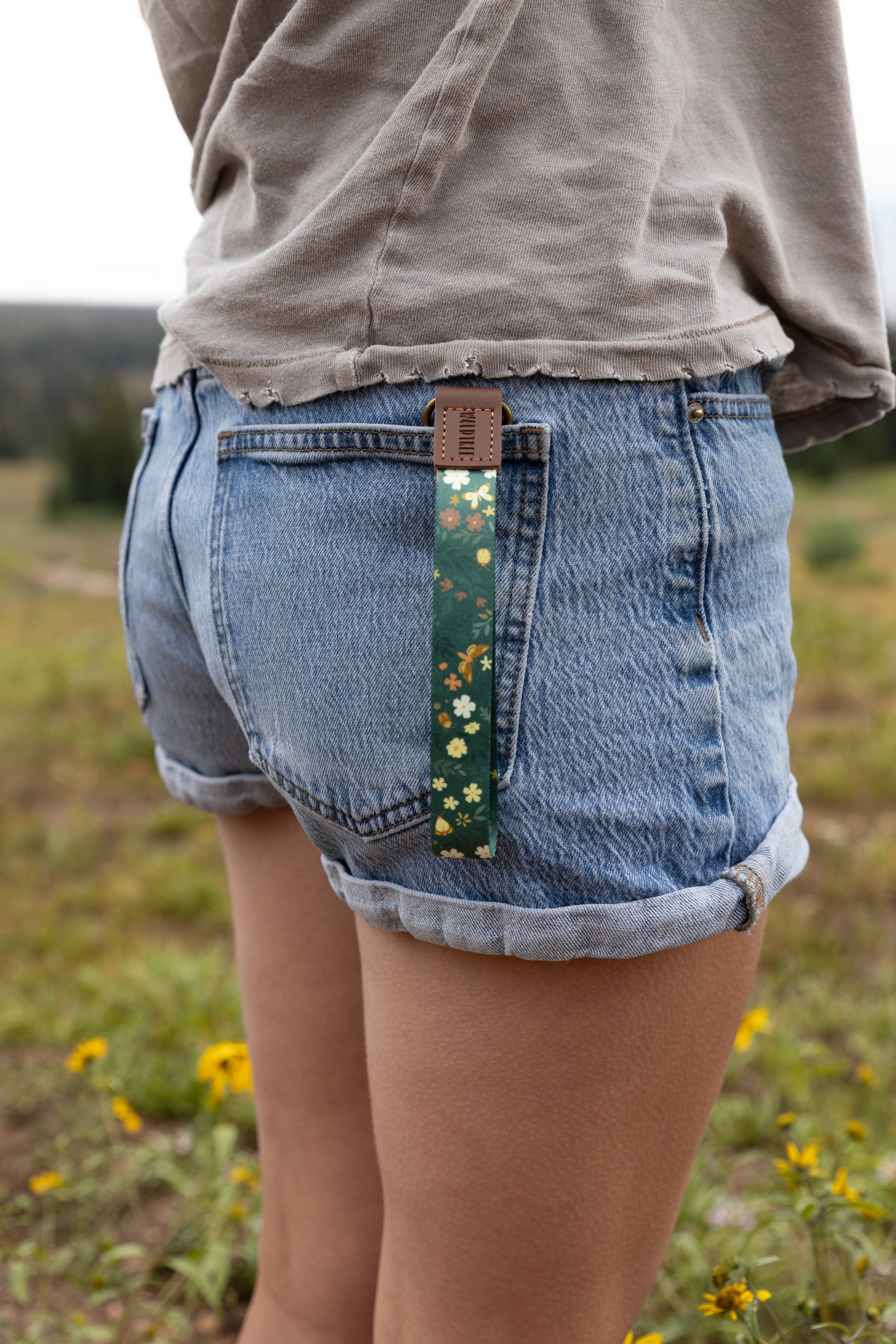 The Flutter & Fern wristlet keychain clipped to the back belt loop of denim shorts, shown from a wider angle in a wildflower meadow, the dark green floral and butterfly print strap hanging against the wearer's leg.