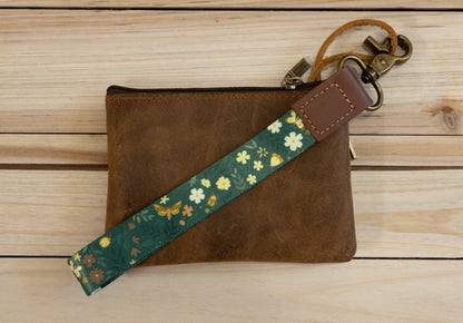 The Flutter & Fern wristlet keychain attached to a set of keys and draped across a brown leather zip pouch on a wood surface, showing the dark green strap printed with white daisies, small flowers, and gold butterflies.