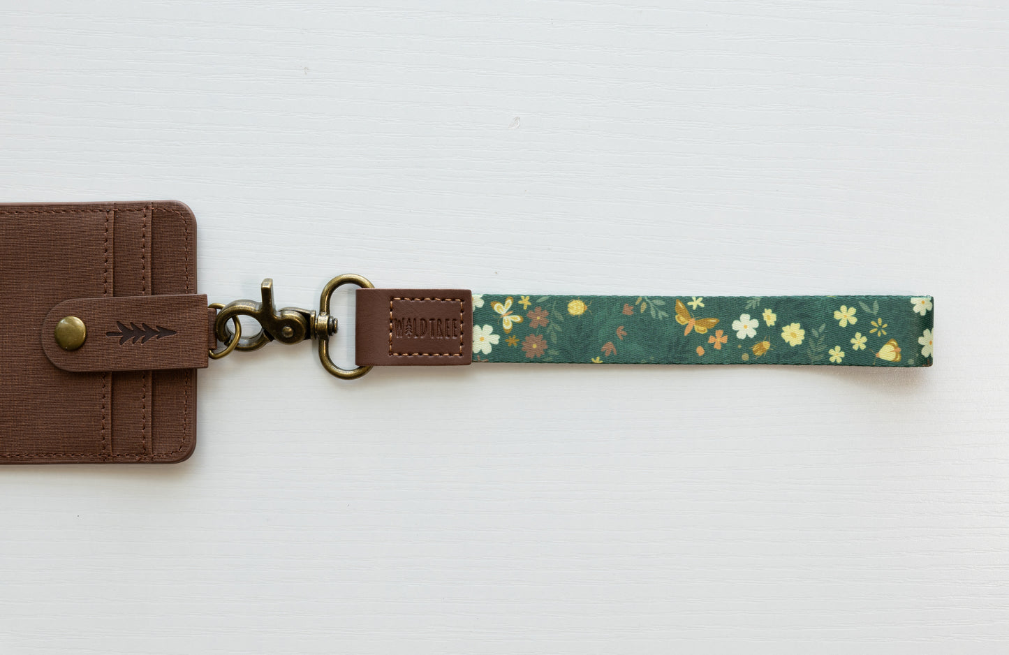 The Flutter & Fern wristlet keychain attached to a brown vegan-leather bifold wallet on a white background, showing the dark green strap printed with white daisies and gold butterflies, the brown leather WILDTREE tab, and bronze swivel clasp.