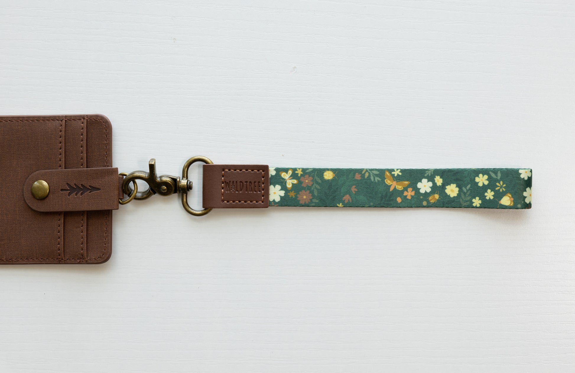 The Flutter & Fern wristlet keychain attached to a brown vegan-leather bifold wallet on a white background, showing the dark green strap printed with white daisies and gold butterflies, the brown leather WILDTREE tab, and bronze swivel clasp.