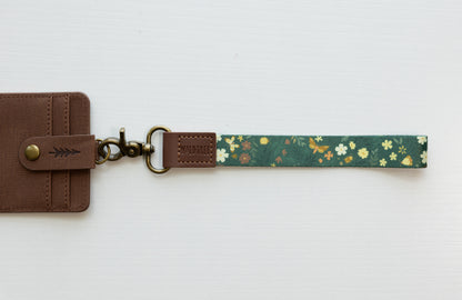 The Flutter & Fern wristlet keychain attached to a brown vegan-leather bifold wallet on a white background, showing the dark green strap printed with white daisies and gold butterflies, the brown leather WILDTREE tab, and bronze swivel clasp.