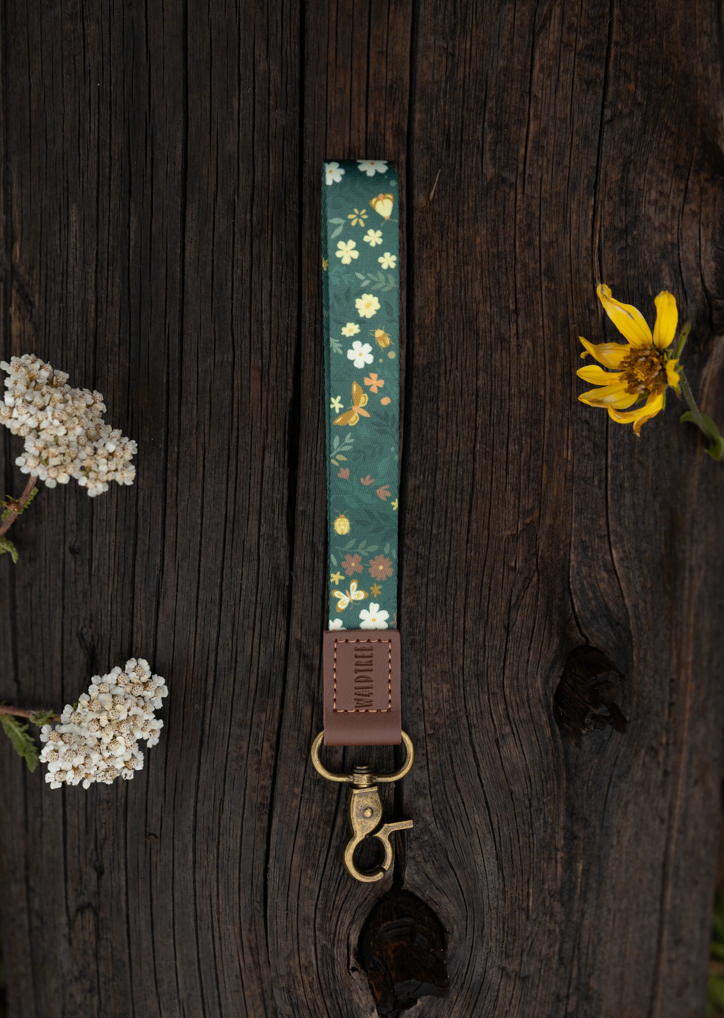The Flutter & Fern wristlet keychain laid on a wooden log surrounded by dried wildflowers, showing the dark green strap printed with white daisies and gold butterflies, the brown leather WILDTREE tab, and the bronze swivel clasp.