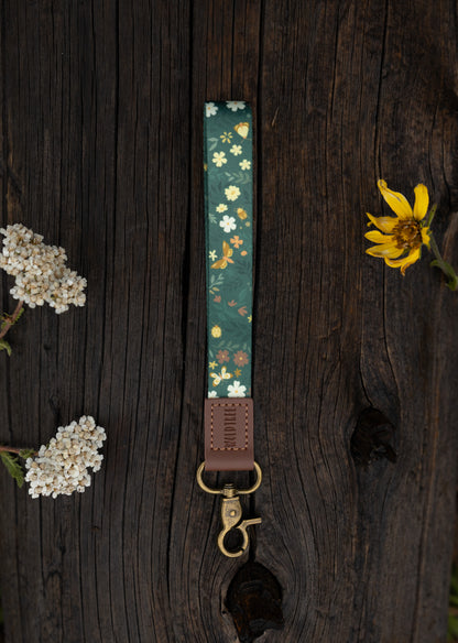 The Flutter & Fern wristlet keychain laid on a wooden log surrounded by dried wildflowers, showing the dark green strap printed with white daisies and gold butterflies, the brown leather WILDTREE tab, and the bronze swivel clasp.
