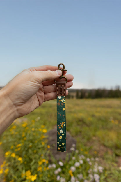 A hand holds the Flutter & Fern wristlet keychain up against a blue sky and wildflower meadow, showing the dark green strap printed with white daisies and gold butterflies, the brown leather WILDTREE tab, and bronze clasps.