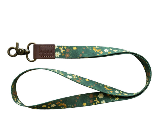 Flat lay of the Flutter & Fern neck lanyard on a white background, showing the full dark green fabric strap printed with white daisies, small brown flowers, and gold butterflies.