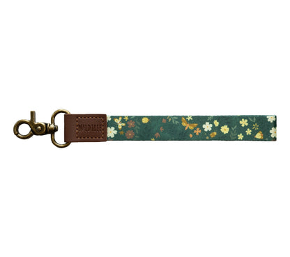 Flat lay of the Flutter & Fern wristlet keychain on a white background, showing the dark green strap printed with white daisies, small brown and yellow flowers, and gold butterflies.