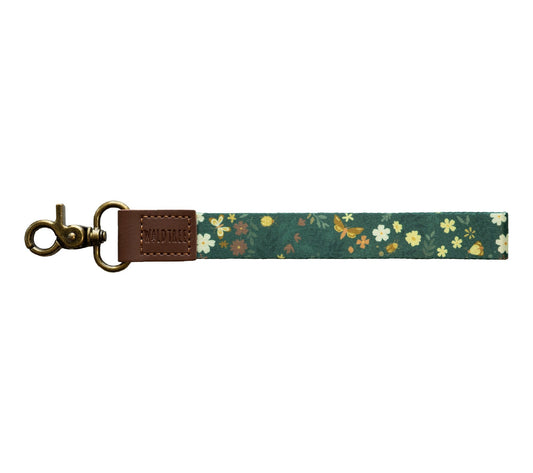 Flat lay of the Flutter & Fern wristlet keychain on a white background, showing the dark green strap printed with white daisies, small brown and yellow flowers, and gold butterflies.