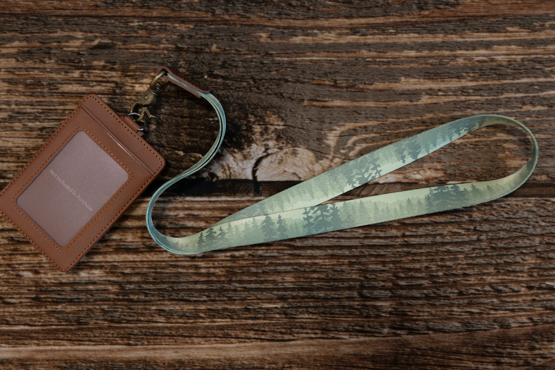 An overhead angled view of the "Wildtree" forest-patterned lanyard paired with a brown leather badge holder on a wooden surface. The badge holder contains a placeholder card that reads "Not Included In Purchase".
