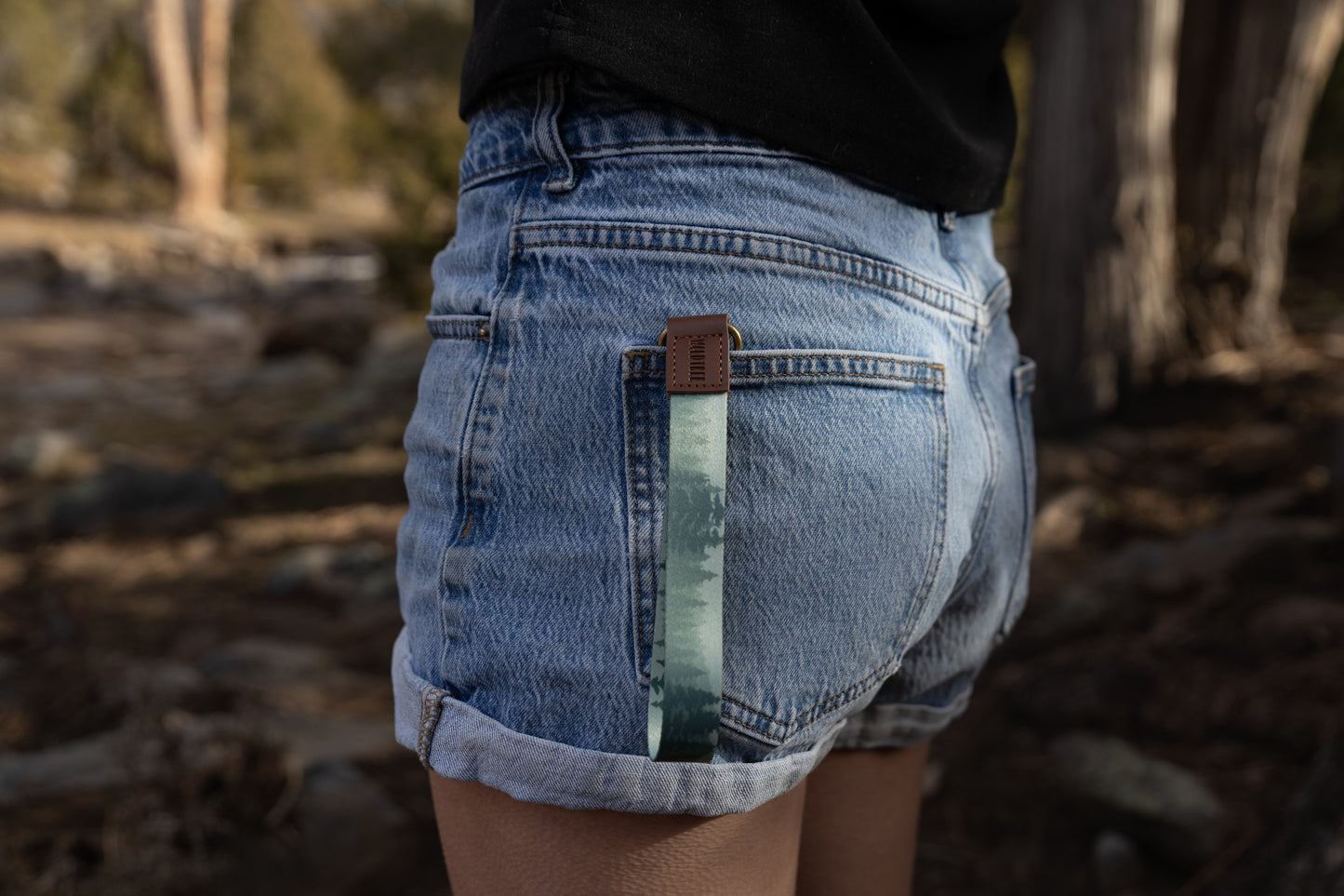 A side-angle shot of a person wearing denim shorts with the foggy forest wristlet keychain tucked into the back pocket. The light green strap hangs down against the denim, highlighting its use as a decorative and functional accessory.