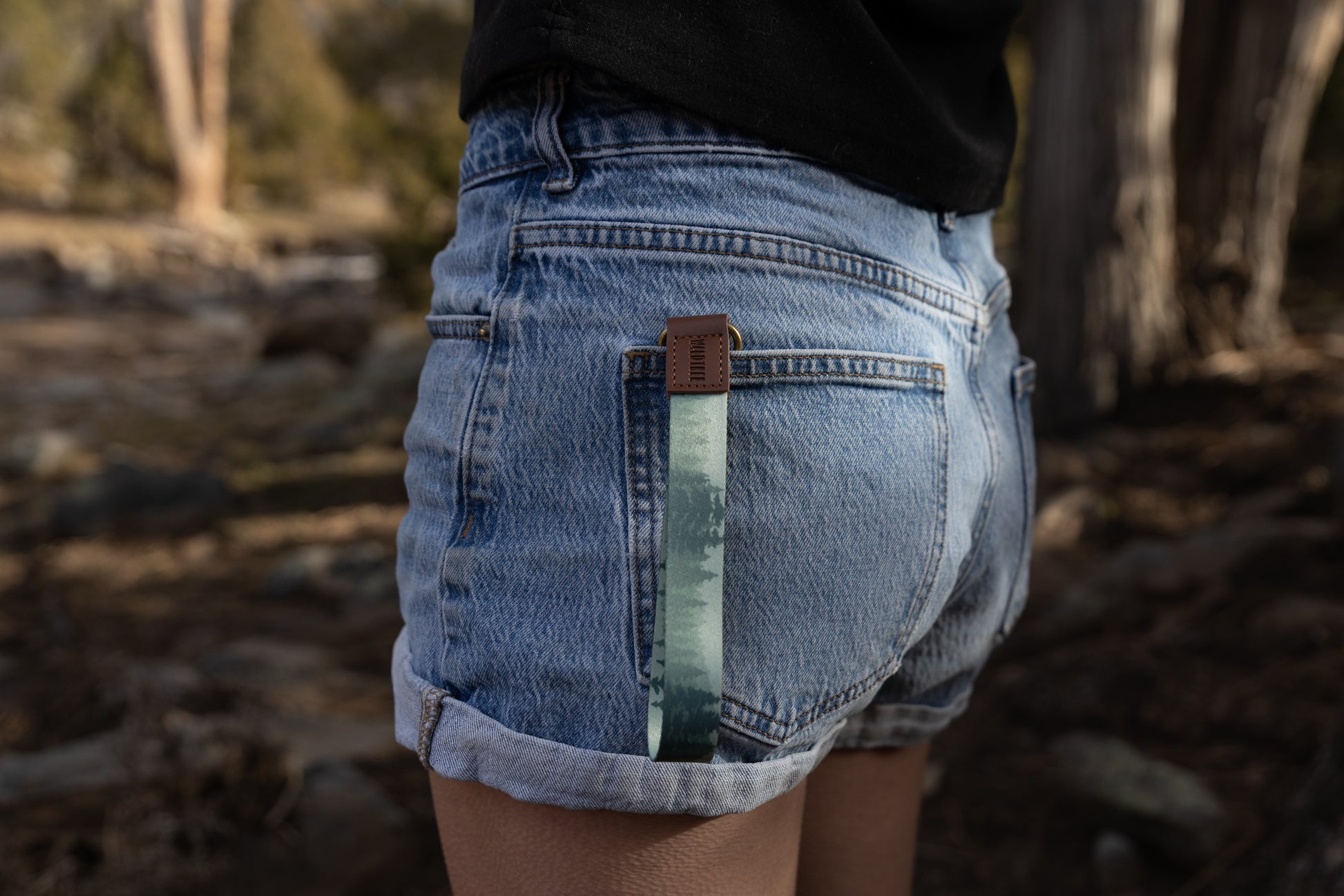 A side-angle shot of a person wearing denim shorts with the foggy forest wristlet keychain tucked into the back pocket. The light green strap hangs down against the denim, highlighting its use as a decorative and functional accessory.