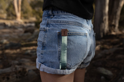 A side-angle shot of a person wearing denim shorts with the foggy forest wristlet keychain tucked into the back pocket. The light green strap hangs down against the denim, highlighting its use as a decorative and functional accessory.